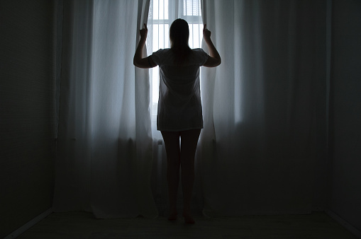 A full-length Caucasian woman standing by the window in the twilight in the morning opens the curtains.