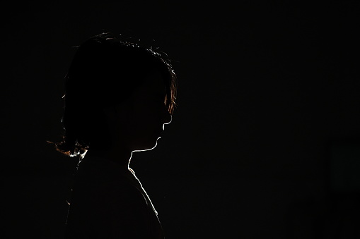 Black foreground and background with backlit silhouette of boy with shoulder length shaggy hair looking straight ahead with hair in face