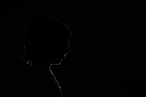 Black foreground and background with backlit silhouette of boy with shoulder length shaggy hair looking down with hair in face