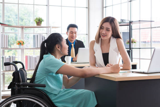 istock Young disabled woman sitting on wheelchair using computer to discuss project with her female colleagues. Business people on wheelchair in the office. Business Woman talking with disabled colleague at desk in modern office. 1300177747