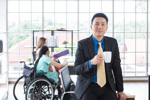 istock Mature businessman standing by the table in his modern office. Confident middle age businessman standing in the office. Handsome CEO smiling, standing in his office with coworkers in the background working by the desk. 1300178199