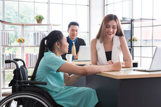 Young disabled woman sitting on wheelchair using computer to discuss project with her female colleagues. Business people on wheelchair in the office. Business Woman talking with disabled colleague at desk in modern office. joven discapacitada sentada en silla de ruedas usando computadora para discutir el proyecto con sus colegas femeninas. gente de negocios en silla de ruedas en la oficina. mujer de negocios hablando con colega discapacitado en el escritorio en la oficina mo