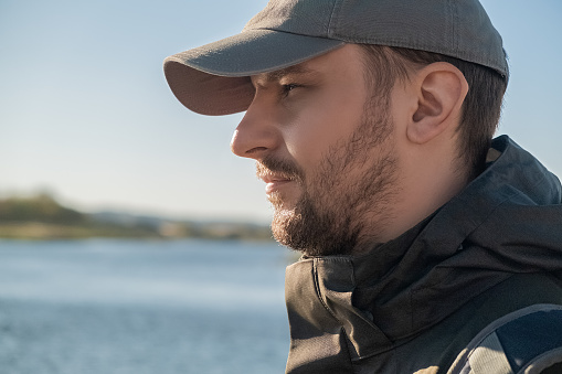 Side view of a serious confident bearded man in a baseball cap looking into the distance on the background of a lake or river on an autumn sunny day