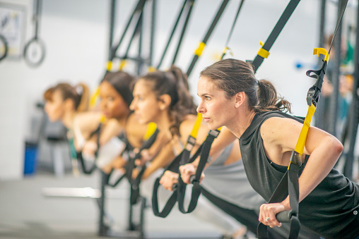 A group of young adults are participating at a group fitness class. They are using suspended resistance bands to test their endurance. A Caucasian woman is in the focus of this photo.