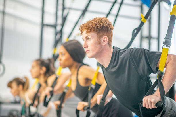 Group fitness class A group of young adults are participating at a group fitness class. They are using suspended resistance bands to test their endurance. A Caucasian male is in the focus of this photo. suspension training stock pictures, royalty-free photos & images