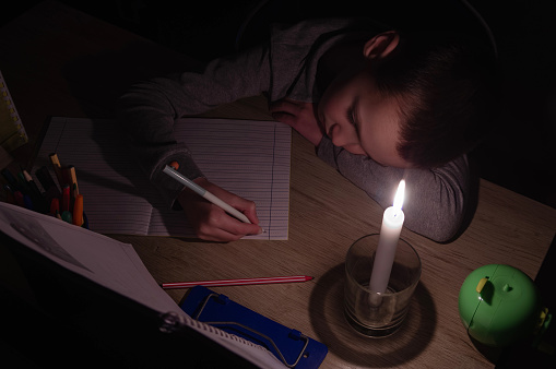Tired schoolboy with candle in complete darkness doing homework. Power outage, blackout, concept image.