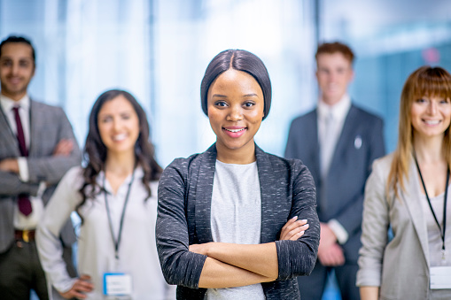 A multi ethnic group of business adults are posing for a photo. A female businesswoman of African  descent is standing at the front of the team and smiling for the camera.