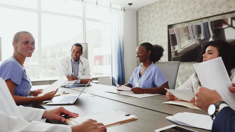 A mature male doctor calls a staff meeting to order. He then hands a document to a female colleague. The female doctor begins to give a presentation. The other healthcare professionals and hospital administrators begin to review copies of the document as the female doctor gives the presentation. The female doctor also uses a laptop during the presentation.
