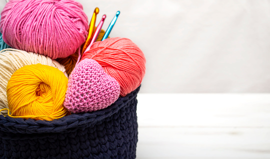 A basket with colored yarn stands on a wooden table, in the basket there is a knitted heart as a symbol of Valentine's Day.
