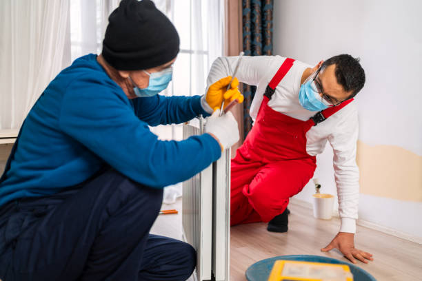 Two professional plumbers with protective face mask are fixing heating radiator with wrench in living room . Heating restoration. Home radiator montage.Rusted radiator