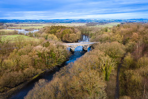 The view from a drone of road bridge crossing a slow moving river on the banks of the river are deciduous trees with bare branches the location is Dumfries and Galloway south west Scotland