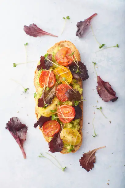 Avocado on toast with cherry tomatoes and salad on a pale background with salad leaves scattered on backdrop