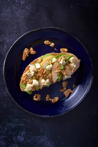 Sliced avocado and pear with gorgonzola and walnuts on toast on a dark blue enamel plate on dark background