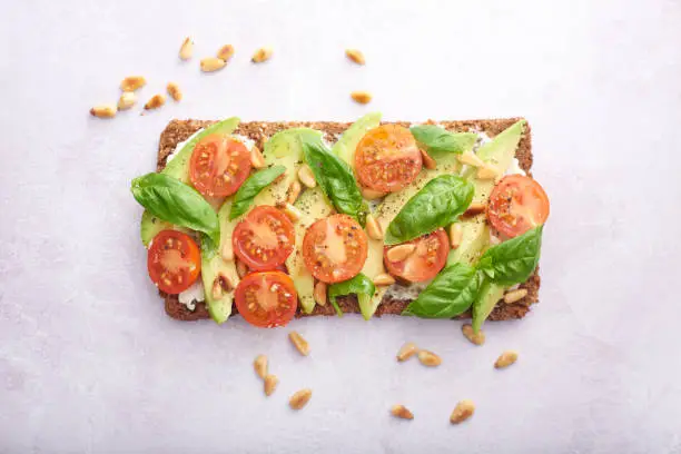 Sliced avocado on rye bread with cherry tomatoes, basil and pine nuts to the side on a pale background