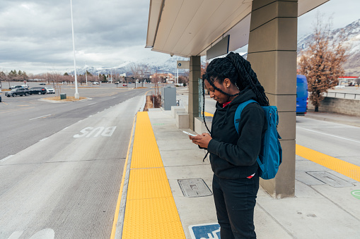 A female college student is at a bus stop waiting for the next bus. She is using public transportation to get to university or college campus and class. Image taken in Utah, USA.