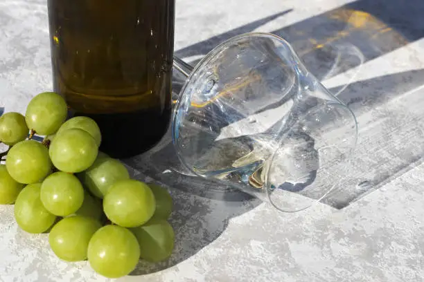 Photo of Wine glass next to a bunch of grapes and a bottle in the sun. Close-up. Place for text. Gray background.