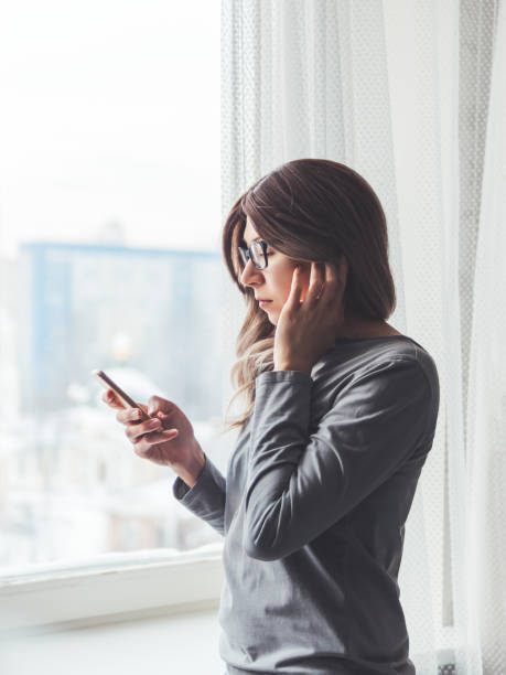 mujer reflexiva con anteojos mira su teléfono inteligente. información en medios en línea. auriculares inalámbricos bajo el pelo. - vertical caucasian glasses red hair fotografías e imágenes de stock