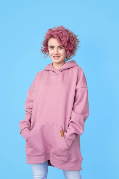 Girl with pink hair in stylish oversized casual hoodie standing and looking at camera Smiling girl with pink hair in stylish oversized casual hoodie standing and looking at camera over blue wall background. Stylish female look, piercing salon, stylist, oversize clothing concept hood clothing stock pictures, royalty-free photos & images