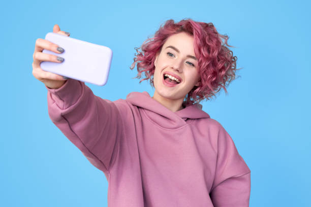 Young pink hair woman making self portrait on smartphone Playful young pink hair woman wearing in trendy hoodie, making self portrait on modern smartphone, standing against blue background with copy space stylish selfie stock pictures, royalty-free photos & images
