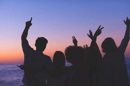 Group of people partying on the beach at sunset or sunrise. They have their arms raised in celebration. Backlit silhouette with copy space