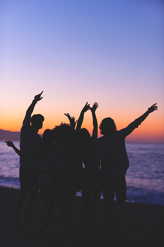 Group of people partying on the beach at sunset or sunrise. They have their arms raised in celebration. Backlit silhouette with copy space