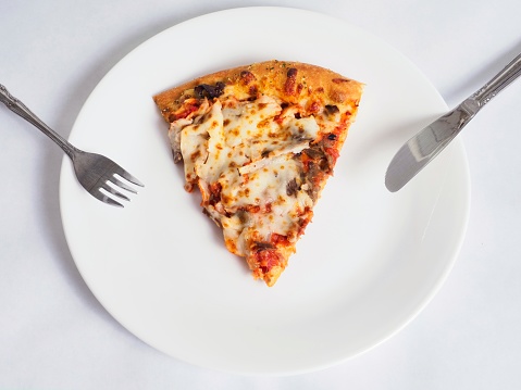 Slice of pizza ready to be eaten with knife and fork on white plate. Messy greasy pizza slice with abundant cheese and sliced beef and onions.