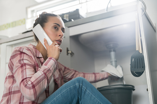 Latin American woman at home calling a plumber about a leaking pipe in her sink and looking very worried