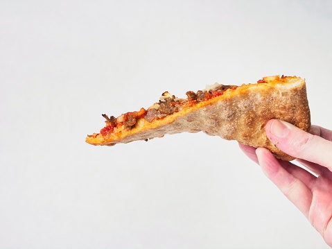 Human hand holding a overloaded slice of pizza folded over and about to fall on the floor. Mozzarella cheese pizza with sliced beef and onions on white background with copy space.