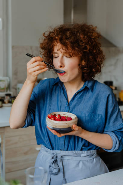 Happy Woman Eating a Bowl of Delicious Oatmeal with Fruit for Breakfast Womam enjoying a healthy breakfast in the kitchen. muesli stock pictures, royalty-free photos & images