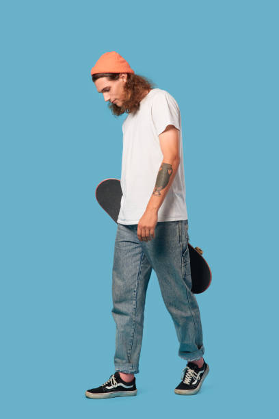 istock Freestyle. Curly-haired guy wearing hat walking isolated on blue with skateboard looking down pensive 1300217918