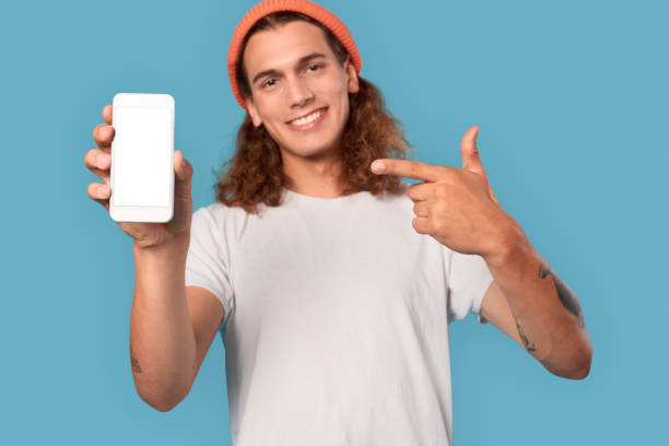 istock Freestyle. Curly-haired guy wearing hat standing isolated on blue pointing at smartphone copy space close-up smiling cheerful blurred background 1300217933