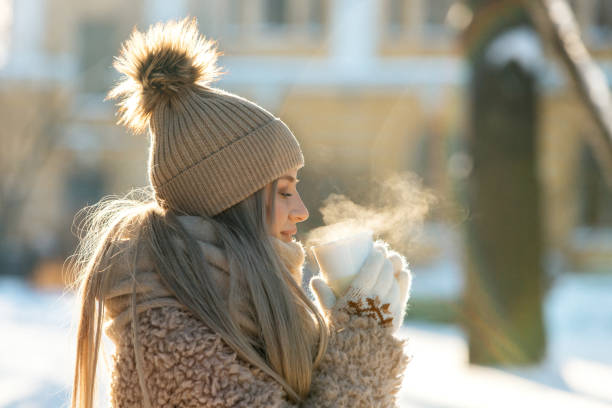 ベージュの毛皮のコートを着た女性、ポンポンの帽子、蒸し白い紅茶/コーヒーを持つミトン、晴れた冬の日 - glove winter wool touching ストックフォトと画像