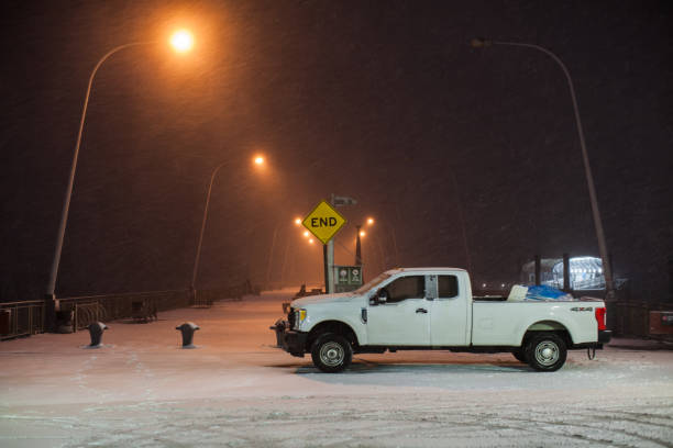 City during bad weather: snowfall and blizzard. White utility worker's car stand at the pier. Yellow road sign \