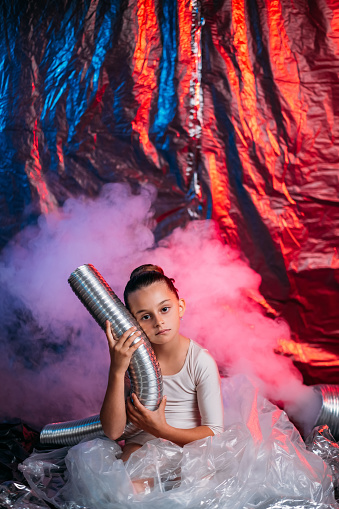 Air pollution. Save nature. Environmental protection. Art portrait of depressed little ballerina girl in polyethylene tutu skirt with pink smoke exhaust pipe on red blue wrinkled foil background.