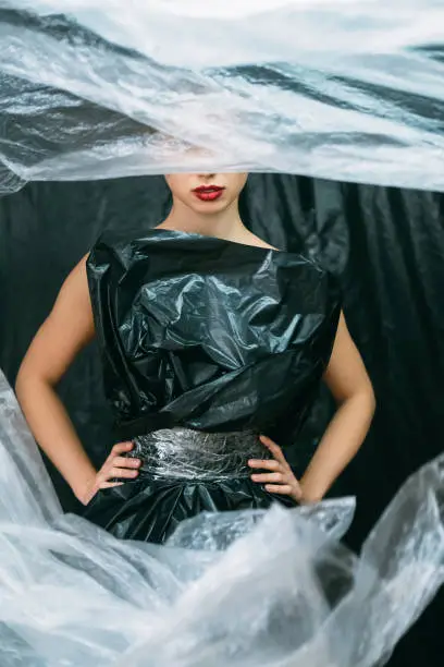 Plastic fashion. Waste reduction. Nature conservation. Air pollution. Female model with red lips posing in black garbage bag dress isolated with white polyethylene film on dark wrinkled background.