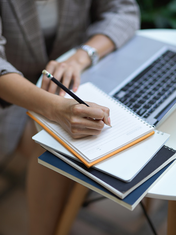 Cropped shot of female hand writing on blank notebook while working laptop on coffee table