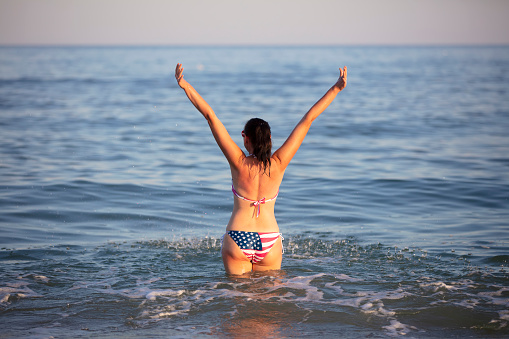 A middle-aged woman stands in sea water with her arms raised in a swimsuit with an American flag print.