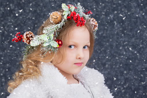 Beautiful little blonde girl with a Christmas wreath on her head on a gray background with falling snow. Christmas child.