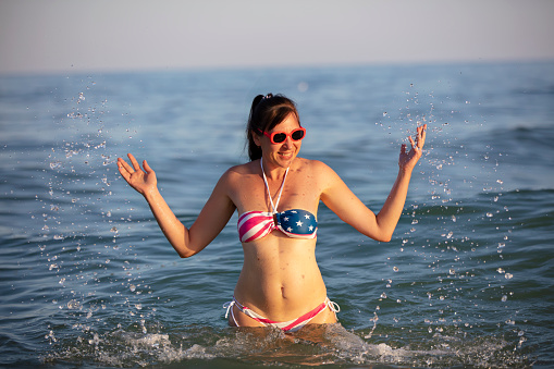Happy middle aged woman splashing sea water in swimsuit with American flag print.