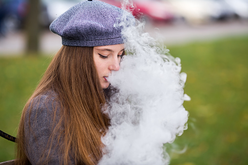 Vaping teenager. Young pretty white caucasian teenage girl in gray beret smoking an electronic cigarette on the bench on the street in the autumn day. Bad habit. Vape activity