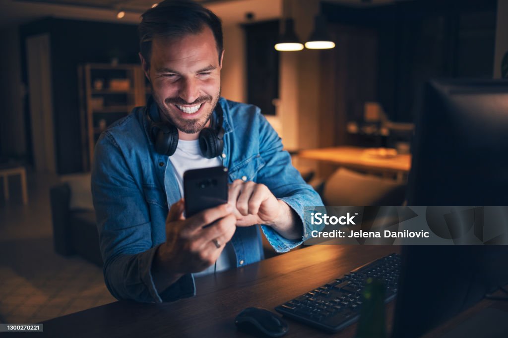 SMS Excited man sitting in his home office, looking at his cellphone with a big smile on face Gambling Stock Photo