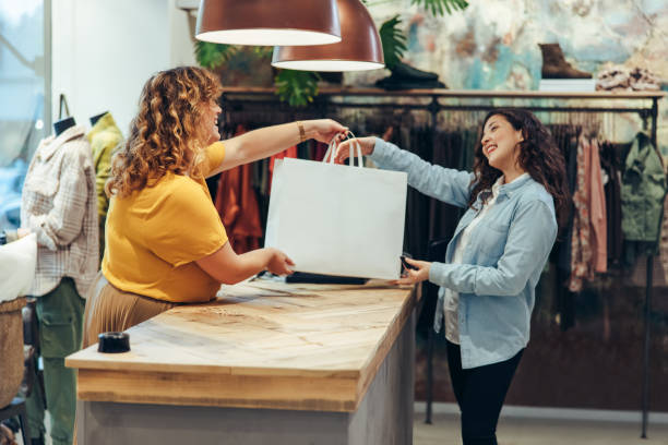 Store owner giving shopping bag to customer Friendly seller of clothing store giving shopping bags to satisfied female customer. Fashion store owner handing over the shopping bag to a female customer at checkout counter. shop stock pictures, royalty-free photos & images