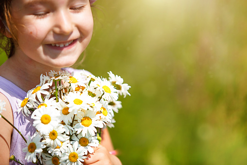 Little girl with a bouquet of daisies in summer on a green natural background. Happy child, hidden face, no face, covered with flowers. Copy space. Authenticity, rural life, eco-friendly