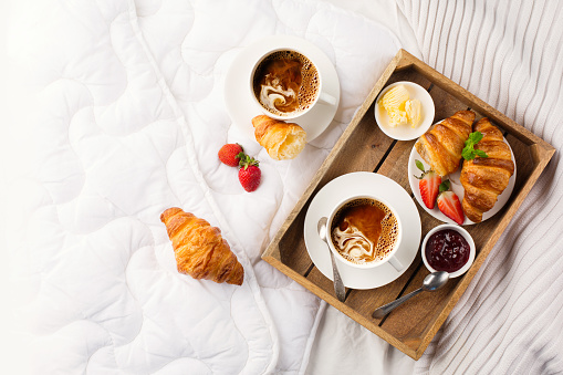 Breakfast in bed, tray with cups of coffee, fresh croissants, jam, fresh strawberries. Honeymoon, valentines day concept. Early morning at hotel. Top view, copy space.