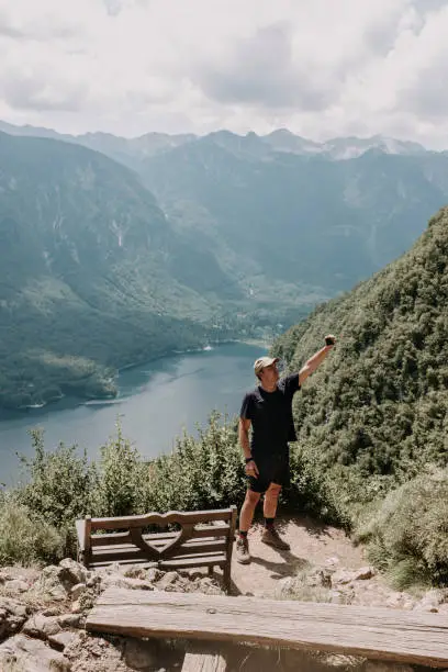 July 29, 2020 - Bohinj, Slovenia: mature man making a selfie with arm stretched standing over the lake Bohinj on the top of the mountains next to bench at the observation point. Hot summer day.