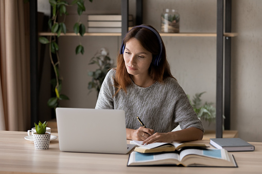 Concentrated millennial Caucasian woman in headphones sit at desk at home study online on laptop. Focused female student in earphones watch webinar take distant education course on computer on web.