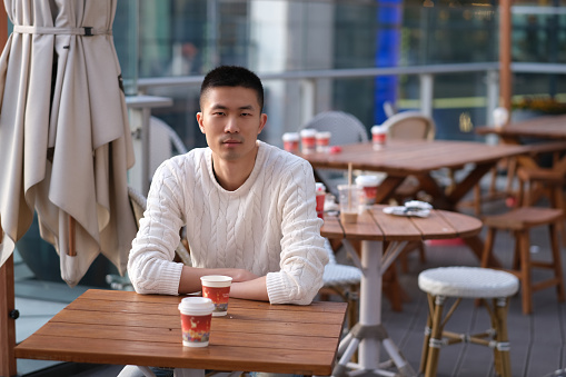 mid shot of one Asian young man in white sweater sitting in open air cafe, looking at camera. blur background