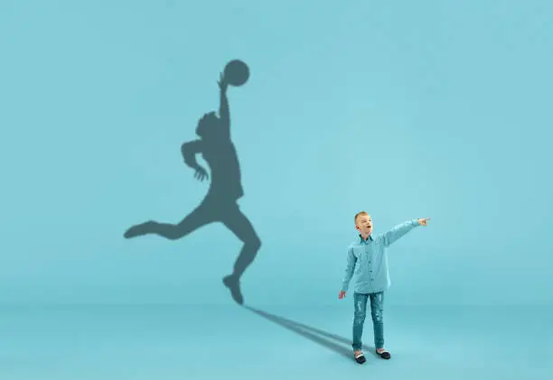 Photo of Childhood and dream about big and famous future. Conceptual image with boy and shadow of fit male basketball player on blue background