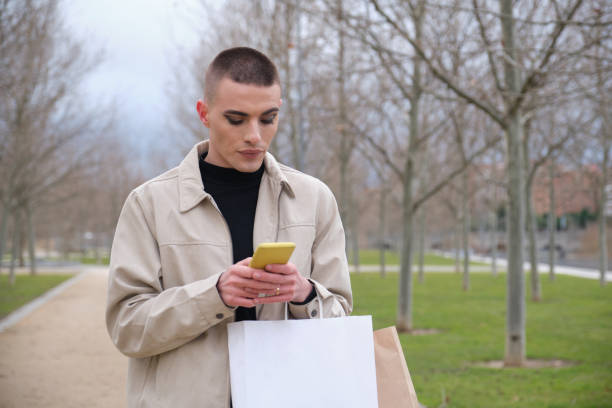 jeune homme beau utilisant le maquillage, bavardant sur son smartphone, transportant des sacs à provisions. type androgyne non binaire. - gay man men male fashion model photos et images de collection