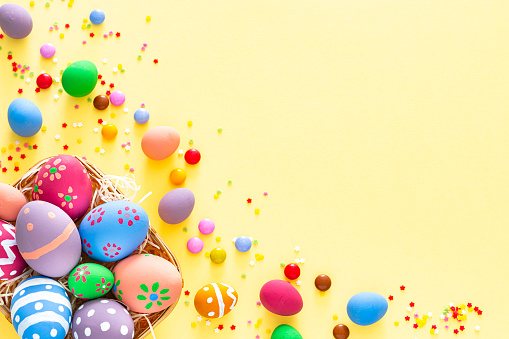 Overhead view of a group of hand painted colorful Easter eggs arranged at the bottom left of a yellow background making a frame and leaving useful copy space for text and/or logo. Sugar sprinkles and candies complete the composition. High resolution 42Mp studio digital capture taken with Sony A7rII and Sony FE 90mm f2.8 macro G OSS lens